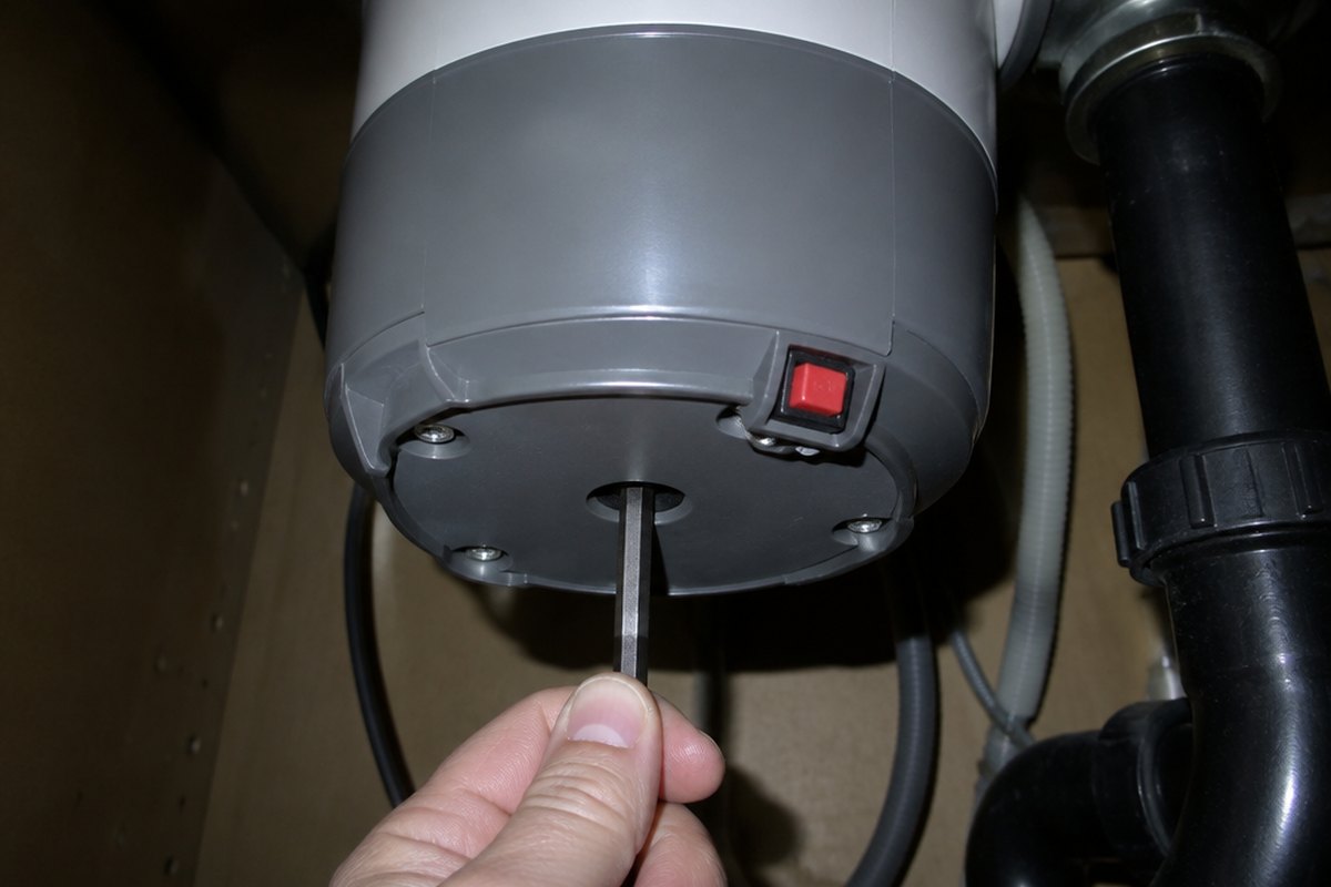 Free a jammed garbage disposal with a hex wrench and the reset button The underside of a garbage disposal with a hex key held in the center socket of the bottom plate and the red reset button on the lower side of the unit